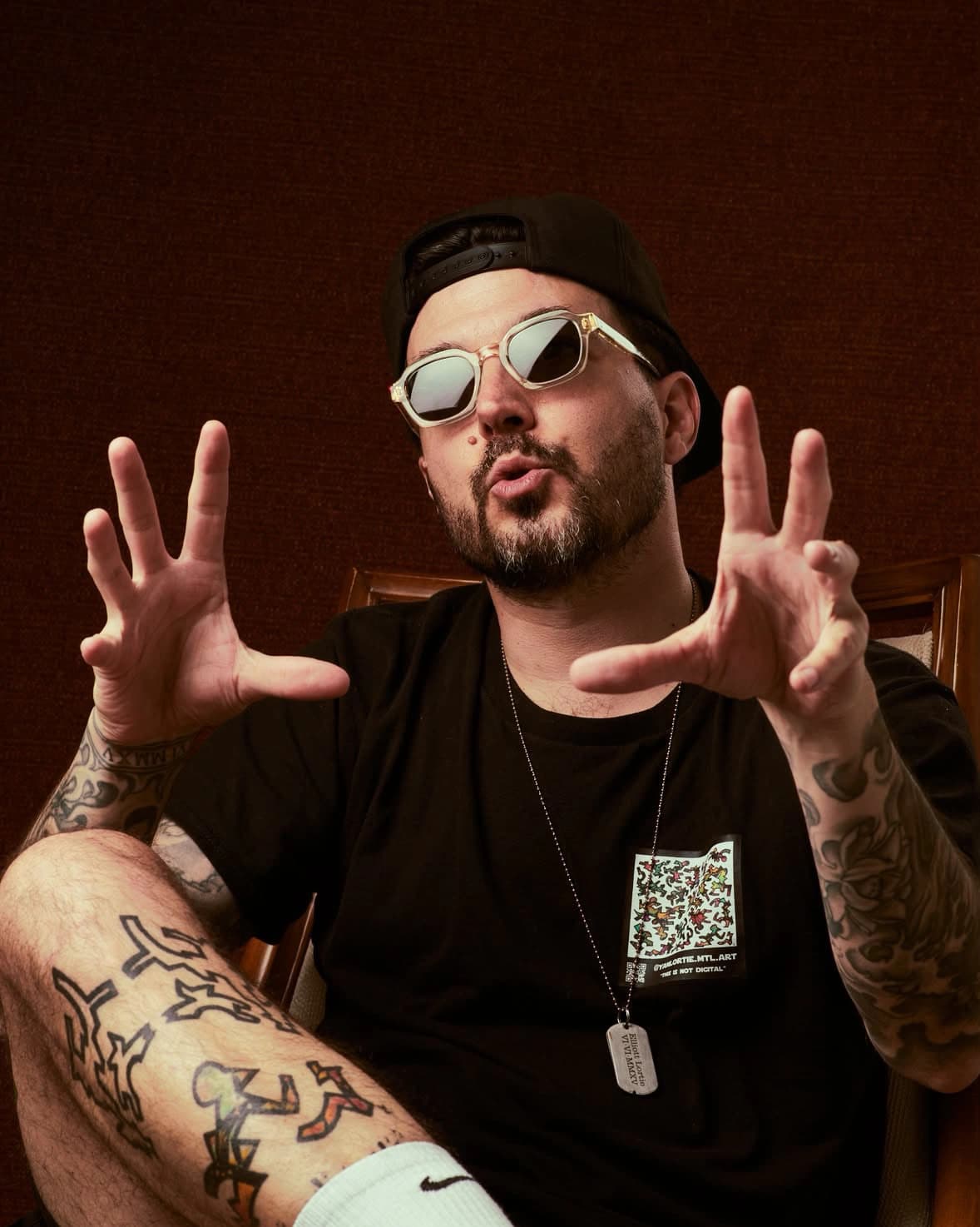 Yan Lortie wearing sunglasses, extending both tattooed hands toward the camera