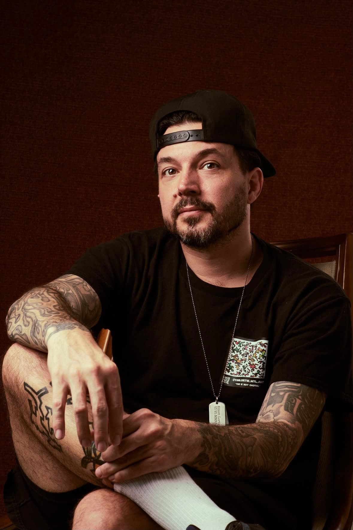 Portrait of Yan Lortie sitting, looking directly at the camera, wearing a backwards cap and a black t-shirt printed with a colourful graphic from his own work, tattoos visible on both forearms